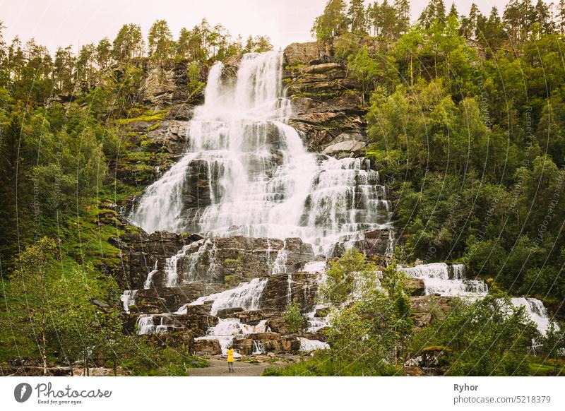 Voss, Hordaland, Norway. Waterfall Tvindefossen In Spring. Largest And Highest Waterfall Of Norway. Famous Natural Norwegian Landmark And Popular Destination. Young Caucasian Woman Lady Tourist Traveler Photographer Taking Pictures Of Waterfall