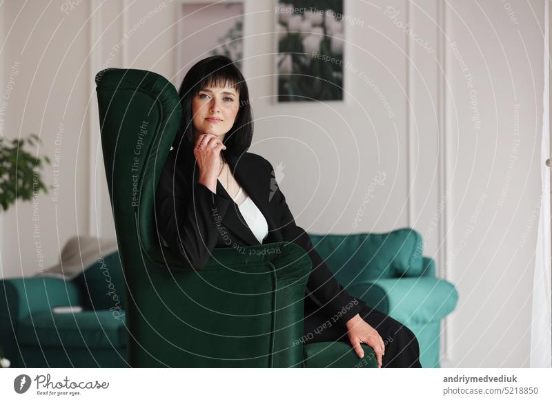 Beautiful business woman in black suit and white blouse is sitting on the green sofa at office, charming brunette female with short hair is looking in camera, girl is working indoors