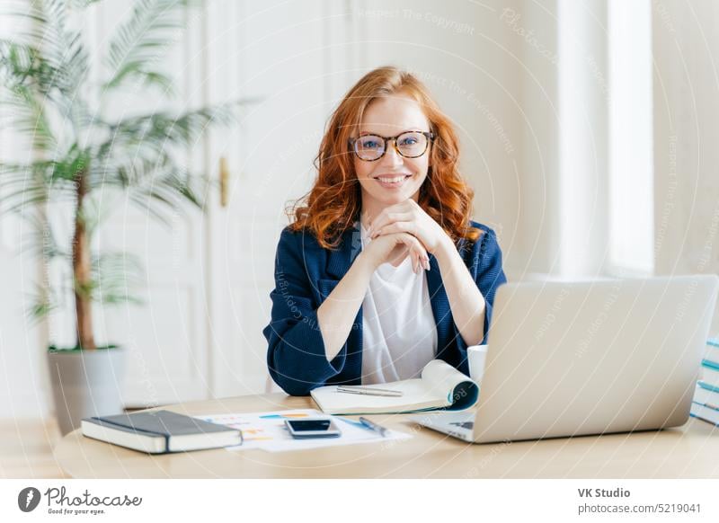 Working concept. Intelligent manager with satisfied expression, writes in notepad, works on modern laptop computer, uses organizer app, sits against office interior. Experienced entrepreneur