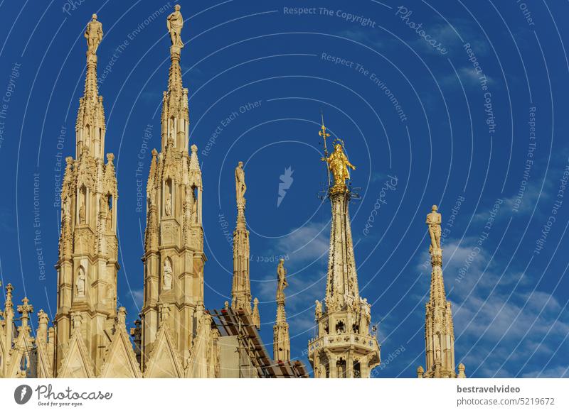 Milan, Italy day view of gothic style Roman Catholic Duomo Cathedral rooftops at homonym main square, with Statue of Madonnina, the Little Madonna, Virgin Mary golden artwork visible against blue sky.