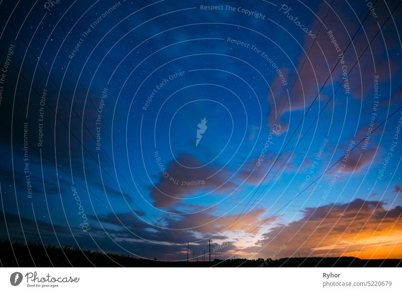 Night Starry Sky With Glowing Stars Above Countryside Landscape. Light Cloudiness Overcast Above Rural Field Meadow In Summer. Power Lines Above Rural Landscape.