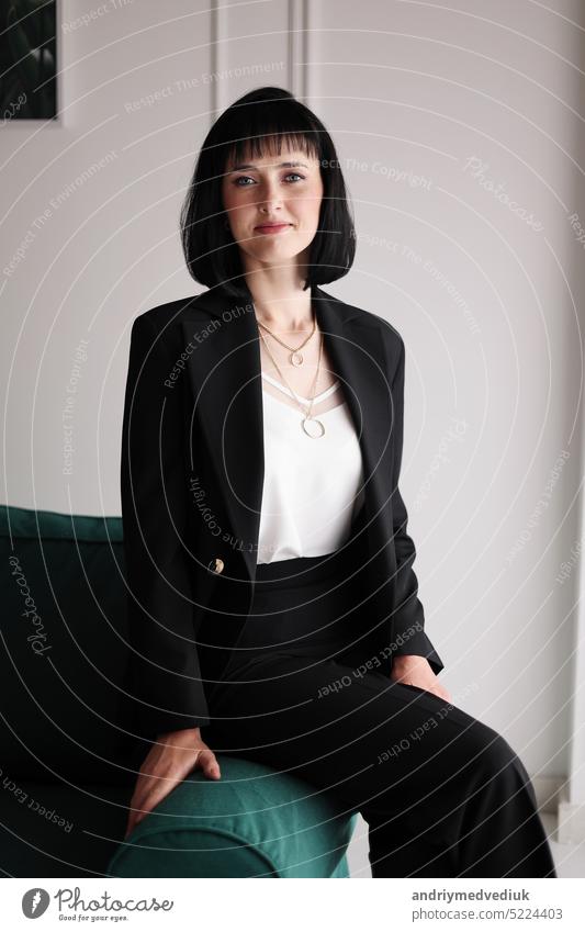 Beautiful business woman in black suit and white blouse is sitting on the green sofa at office, charming brunette female with short hair is looking in camera, girl is working indoors