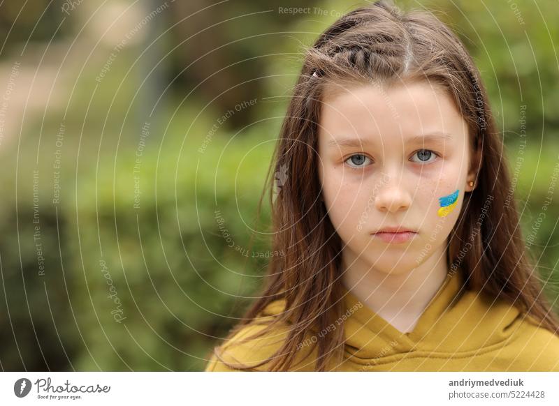 the face of a frightened girl, painted on her cheek in the yellow-blue colors of the Ukrainian flag, a request for help. Children ask for peace. High quality photo. High quality photo