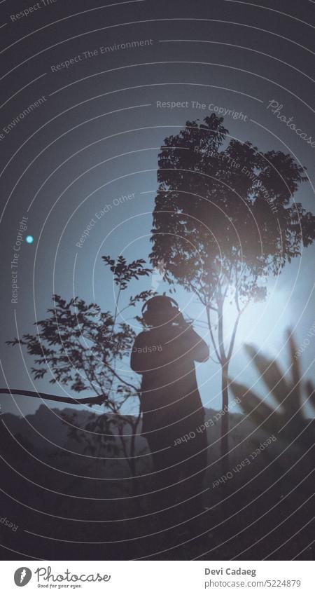 A silhouette of a woman with headphones taking picture under the sun Silhouette Sky Tree Blue Branch Tree trunk Sun Nature Clouds Summer Green Camera