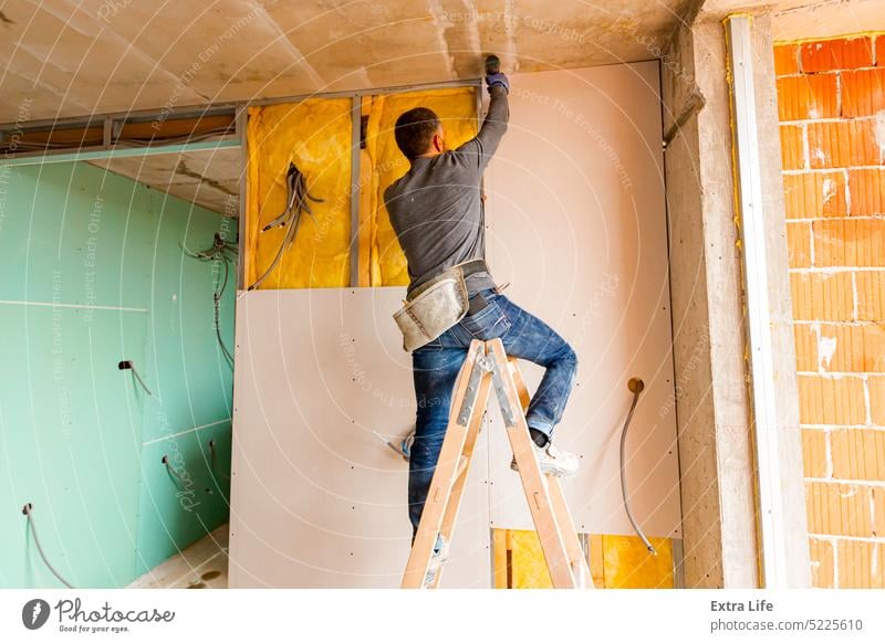 Worker on ladder uses hand drill, ACU power tool and twists screws on rock wool isolated gypsum dry wall Assembly Battery Block Board Bolt Brick Building Site