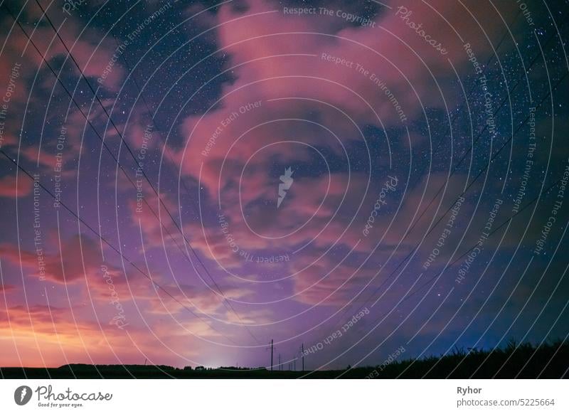 Night Starry Sky With Glowing Stars Above Countryside Landscape. Light Cloudiness Overcast Above Rural Field Meadow In Summer. Power Lines Above Rural Landscape