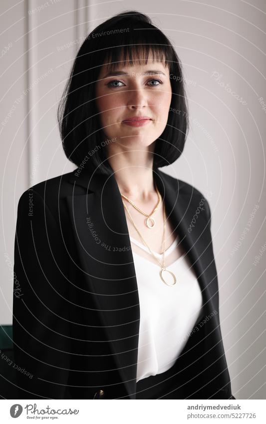 Beautiful business woman in black suit and white blouse is sitting on the green sofa at office, charming brunette female with short hair is looking in camera, girl is working indoors