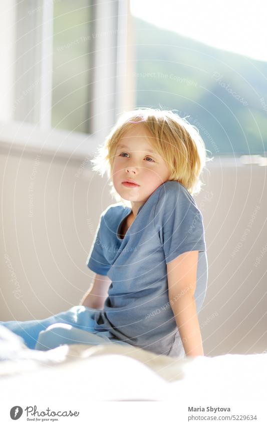 Vertical portrait of cute dreamy schoolboy child in a blue tshirt in the bedroom on sunny day. Preteen boy sitting on the bed and dreaming. preteen haircut