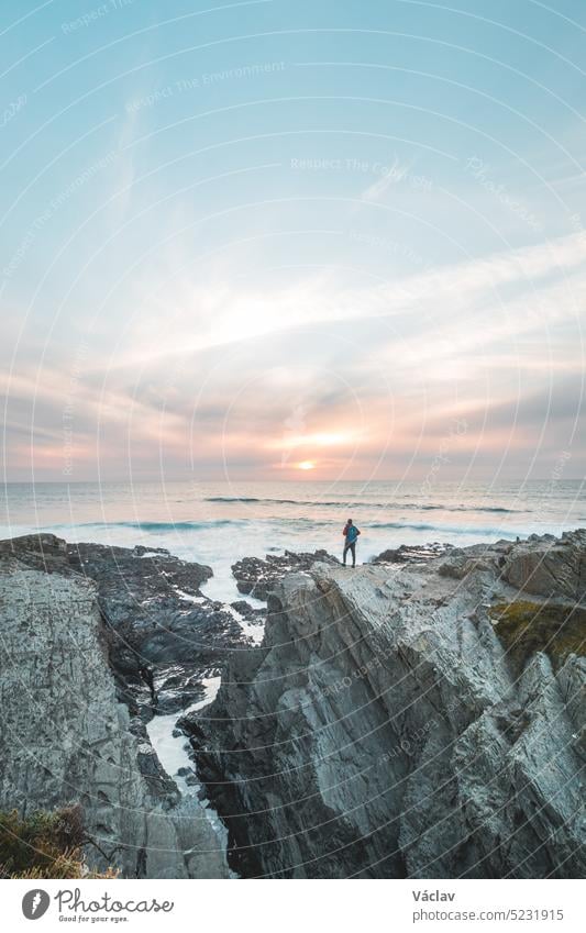 Fearless man with a backpack watching the sunset over the Atlantic Ocean in Porto Covo, Portugal. A hiker stands on the edge of a cliff looking into his future. Self-awareness