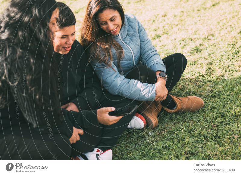 Hispanic family spending quality time together in a local park on a beautiful, sunny day. A teenage boy is sitting on the grass, holding his smartphone while looking away. His mother and sister are sitting next to him, looking at the camera and smiling.