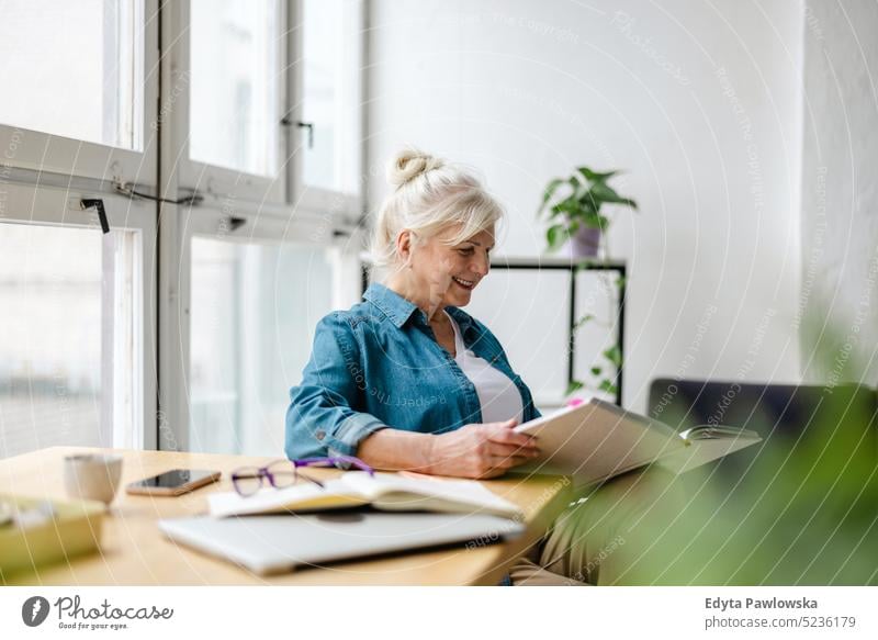 Smiling mature businesswoman writing in notebook while sitting at table in office real people senior indoors loft window home mature adult one person attractive