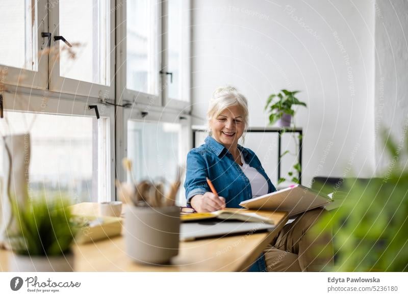 Smiling mature businesswoman writing in notebook while sitting at table in office real people senior indoors loft window home mature adult one person attractive