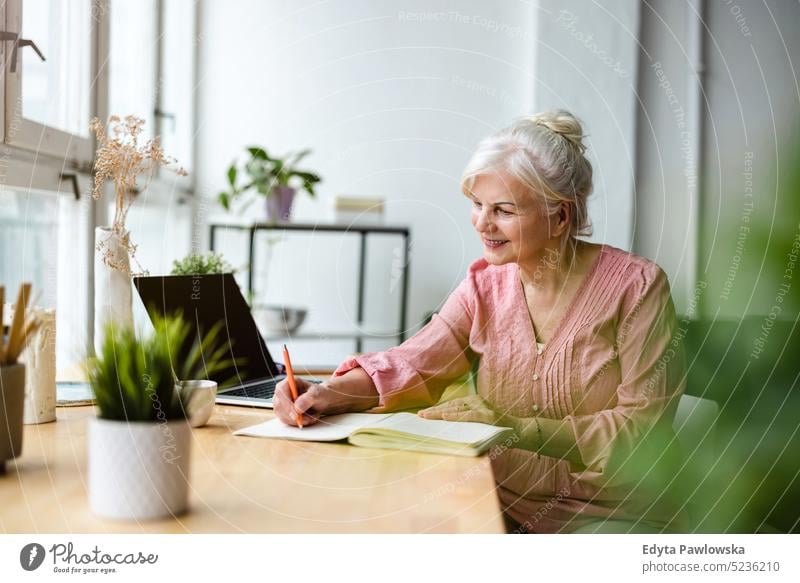 Smiling mature businesswoman writing in notebook while sitting at table in office real people senior indoors loft window home mature adult one person attractive