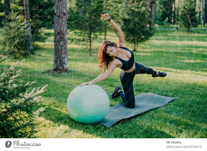 Flexible active woman in sportsclothes makes fitness exercises on karemat with fitball, raises arms and breathes fresh air in forest has morning workout poses over nature background. Healthy lifestyle