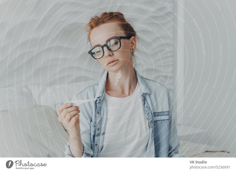 Young unhealthy red-haired lady holding thermometer while sitting on bed at home woman measuring temperature upset young suffering flu influenza ill morning