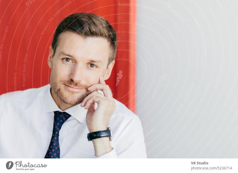 Self assured unshaven male office worker in formal white shirt with tie, holds hand on cheek, thinks about starting new stage in life, poses against red and white background. Business concept