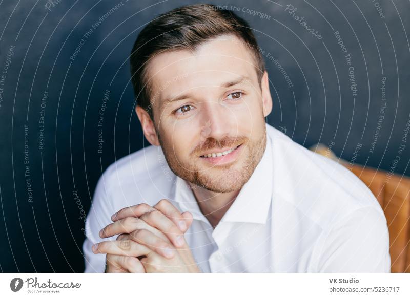 Photo of cheerful pensive young male with bristle, dreams about something pleasant, wears white shirt, poses against dark wall. Successful prosperous male executive worker contemplates indoor