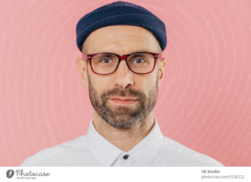 Close up shot of confident male employee with thick beard and mustache, wears transparent glasses, hat, white shirt, stands against pink background, has attentive gaze at camera. Facial expressions