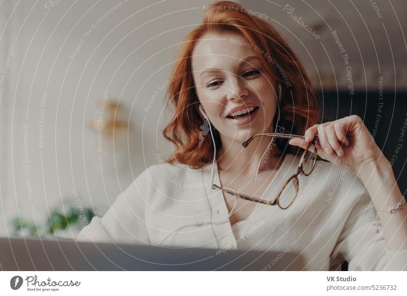 Confident pleased redhead woman concentrated in laptop computer, watches video or movie on laptop computer, enjoys friendly video call, holds transparent glasses, uses free internet connection