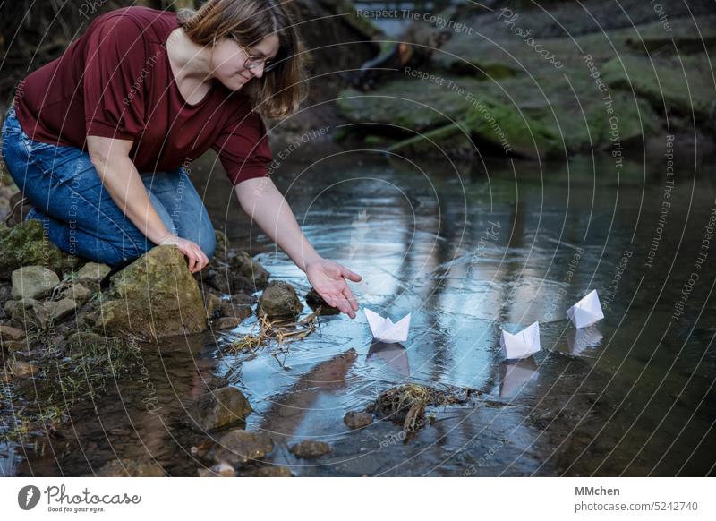 Woman sits on shore and drops paper boat into water Water Paper boat bank Good mood Stony voyage Mystic Ice vacation Cruise Happiness Joie de vivre (Vitality)