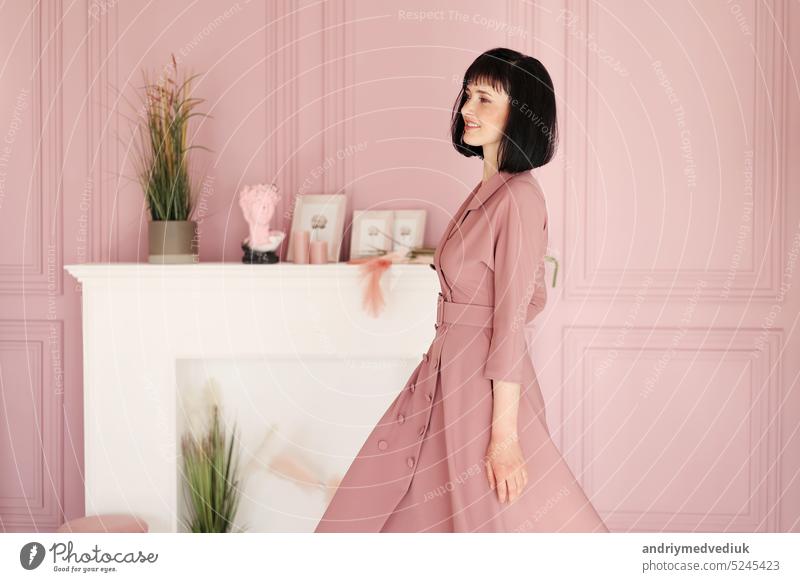 Portrait of attractive smiling brunette with short hair woman folded arms staying alone at stylish interior indoors. Female in soft pink dress is looking in camera