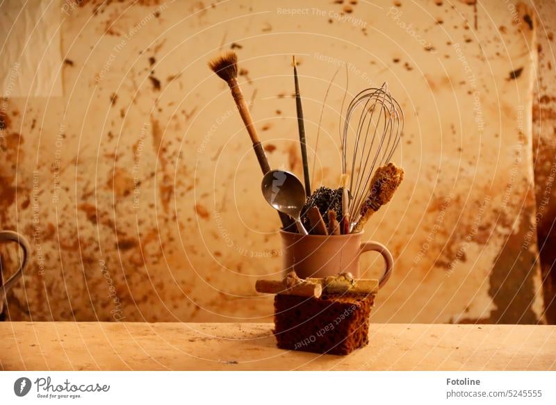 On an old table is a cup with various tools in it. in front of the cup is a sponge. The wall in the background is dirty. No one has worked here for a long time.