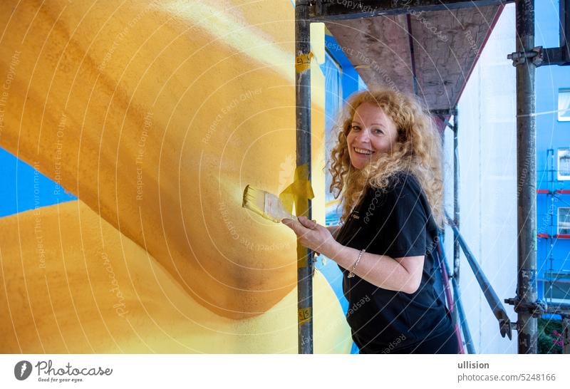 Redhead female smiling, laughing artist painter woman with curly red hair in her prime stands on a facade scaffolding and paints a sunflower mural on the house wall