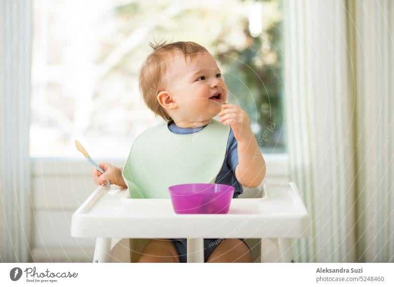 Cute baby eating first solid food, infant sitting in high chair. Child tasting vegetables at the table, discovering new food. Cozy kitchen interior. Healthy food concept.