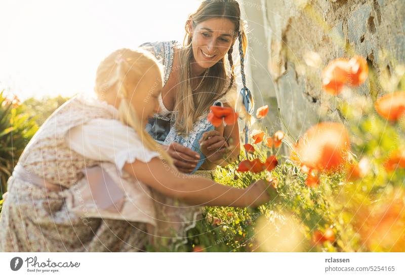 Sweet little girl and her mother in traditional bavarian tracht picking poppies plants outdoor in a village at oktoberfest house young summer nature landscape