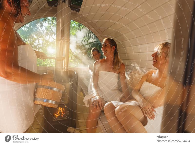 Wellness - Happy friends have fun at the wooden barrel sauna, water and scent are splashed on hot stones for steam in norway relaxing in the finnish sauna cabin.