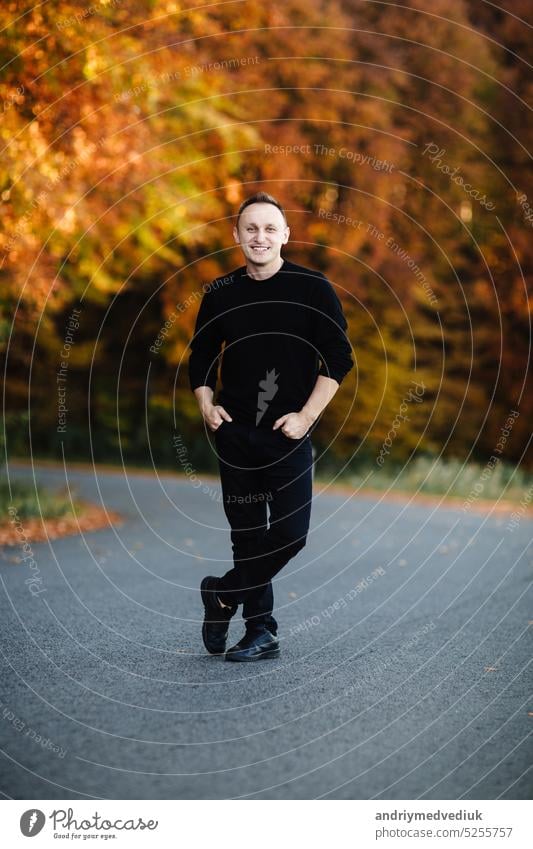 Young attractive man is standing on road in autumn forest. Guy with short hair wearing in black is looking in camera in park with yellow leaves trees. Cozy day on countryside