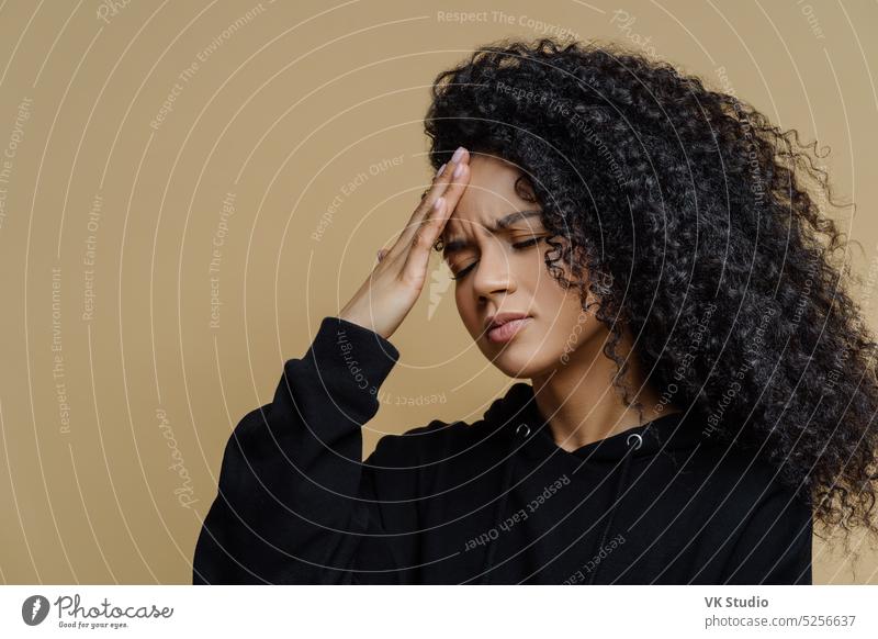 Sideways shot of displeased dark skinned woman touches forehead, suffers headache and mirgaine, squints face from pain, needs painkillers, cannot concentrate, wears black hoodie stands over beige wall