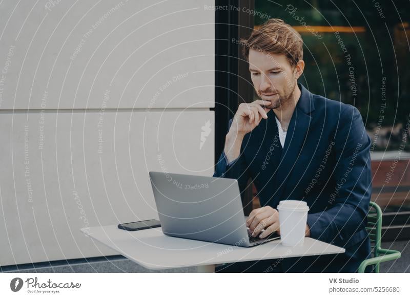 Handsome pensive young man browsing web on laptop while working remotely in sidewalk cafe thoughtful businessman coffee table outdoors outside distant online