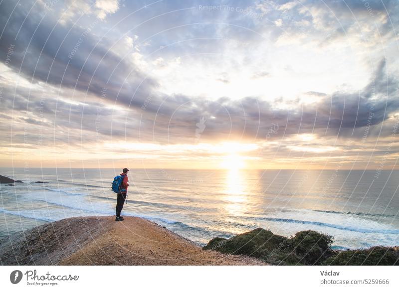 Traveler stands on the edge of a cliff near the town of Odeceixe, Algarve region, Portugal watching the sun setting on the clear blue ocean. The play of colors in the sky