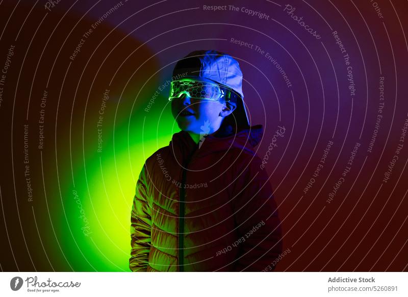 Child in futuristic glasses under neon illumination child outerwear dark illuminate smile goggles happy style content glow cap glad positive childhood club coat