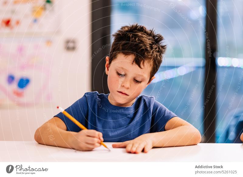 Boy sitting at table and drawing boy home window creative hobby evening child glad childhood kid pencil sketch cute imagination casual dark hair adorable desk