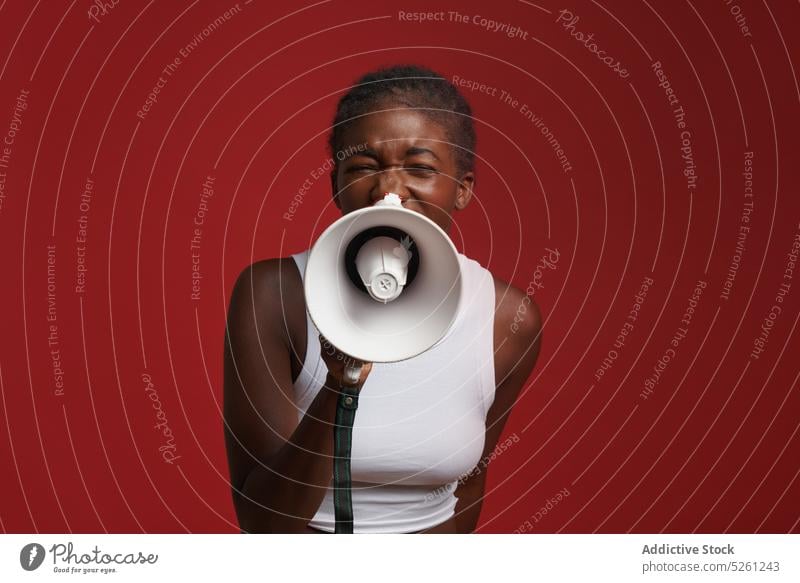 Ethnic woman with megaphone near red wall shout loudspeaker braid scream announce studio sportswear hairstyle eyes closed female african american ethnic black