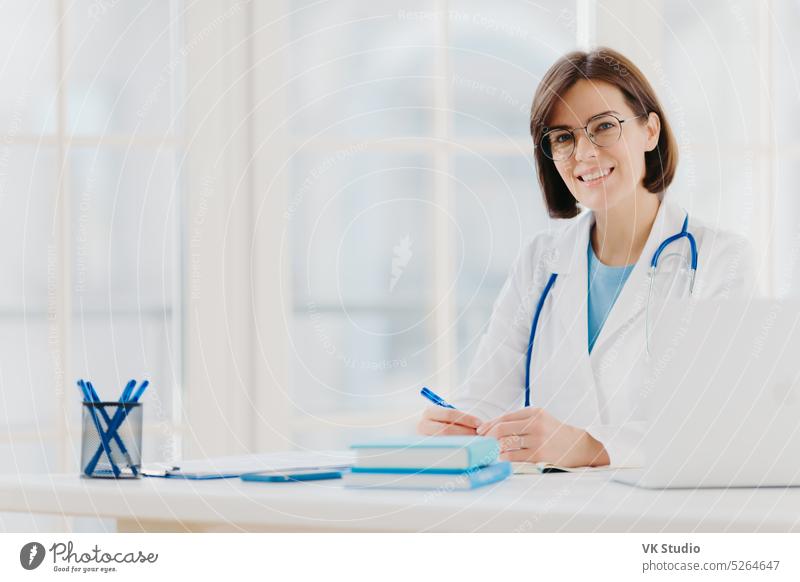 Medical specialist wears white gown with stethoscope, works in hospital, makes notes, poses at own cabinet, sits at table with laptop computer in office smiles happily. Health care and medical concept