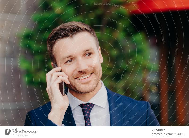 Headshot of handsome unshaven young male has conversation with service operator, has happy expression, attractive look, dressed in formal outfit, poses outdoor. People and lifestyle concept.