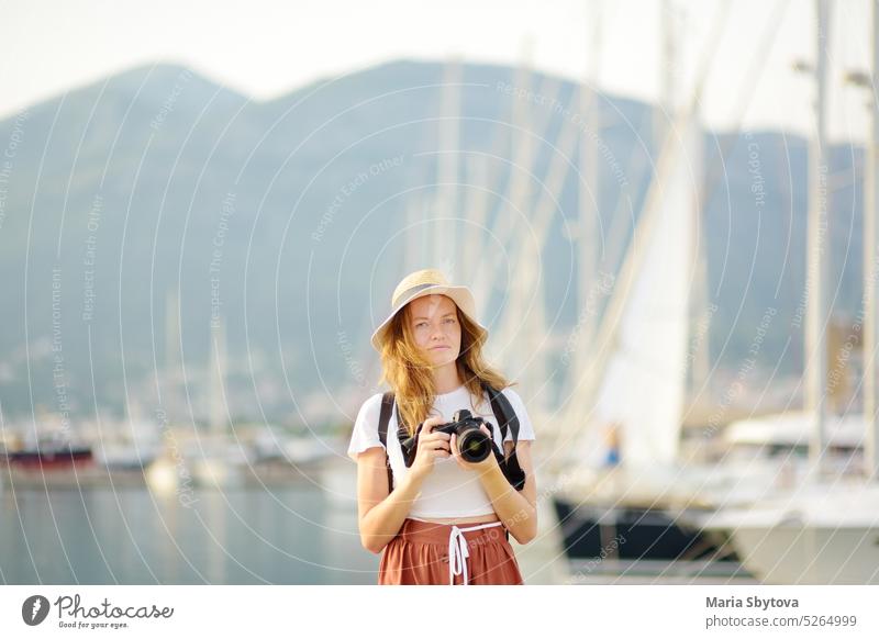 Charming young tourist taking a photo on the Mediterranean coast. Attractive redhead girl photographer with a camera on the background of sea and yachts. Tourism and travel