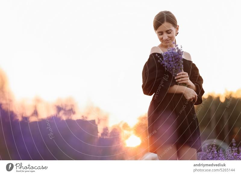 Young romantic woman with healthy natural beauty skin holding bouquet in lavender field. Beautiful girl wearing black dress and bow on hair. Advertising natural product