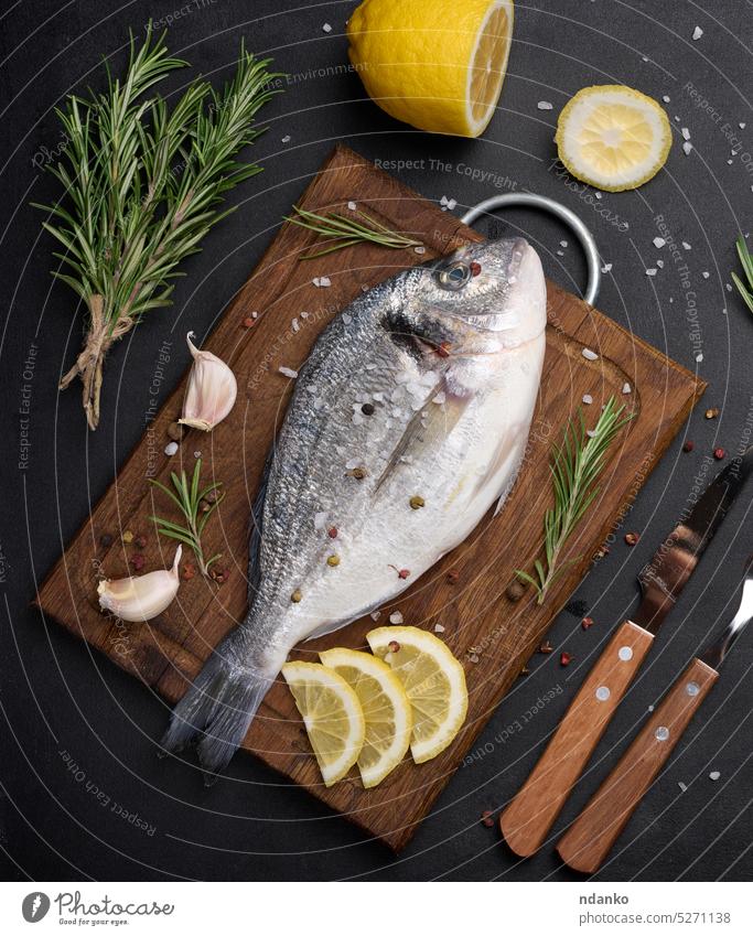 Raw whole dorado fish on wooden kitchen board and spices for cooking, top view on black table raw seafood rosemary salt lemon garlic preparation freshness