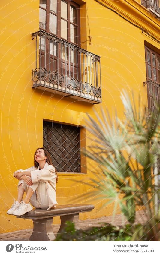 Young woman sitting on the bench, smiling, near yellow building with beautiful balcony. Female tourist traveling solo. millennial urban model fashion wall city