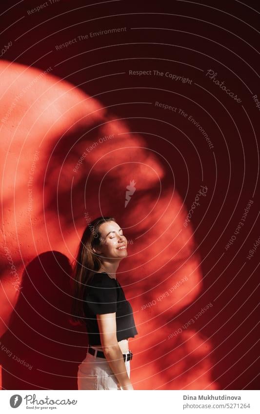 Young woman standing near red wall smiling. Artistic photo with female model and shadows on the wall. millennial urban fashion city tourist tourism young adult