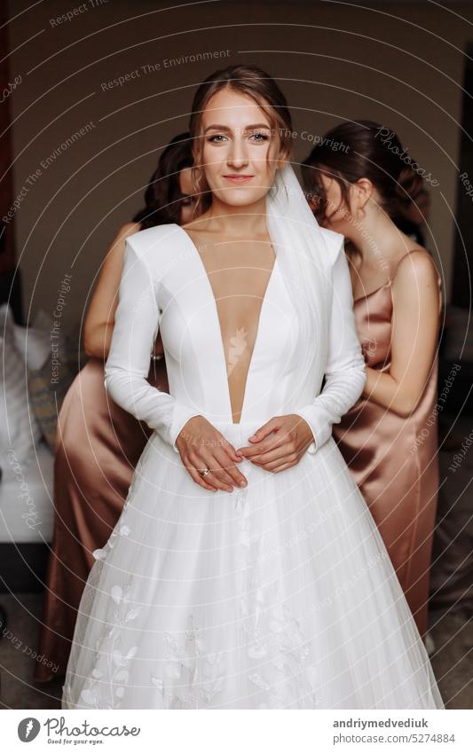 beautiful smiling young bride in long white satin dress with deep neckline, hairstyle and make up standing in hotel room. bridesmaids help her preparation for the wedding day in the morning