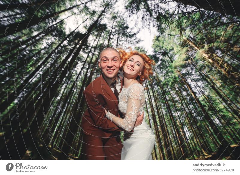 Happy stylish couple newlyweds in the green forest on summer day. bride in long white dress and groom in red suit are hugging. wedding day. happy park nature