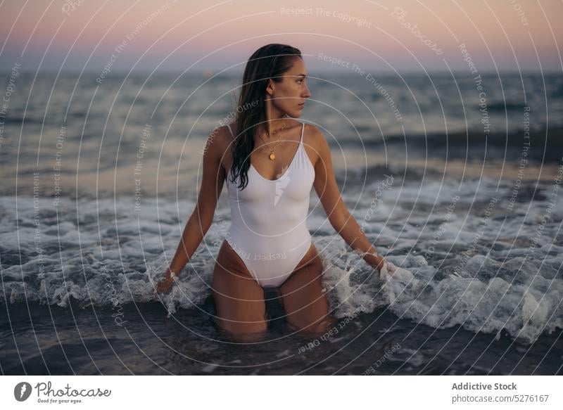 Young woman sitting in sea waves beach sunset kneel summer weekend resort water female young dark hair shore ocean evening twilight vacation swimsuit coast