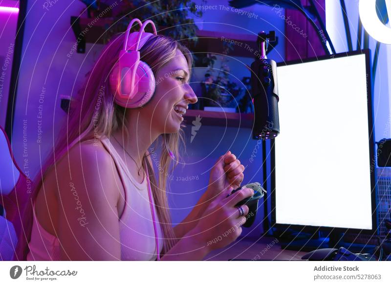 Cheerful female streamer in headphones using mic and gamepad woman cheerful console play happy microphone record blogger neon room headset talk young illuminate