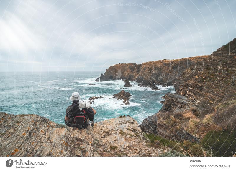 Hiker and adventurer sit on a rugged rock and cliff coastline on the Atlantic Ocean near the town of Odeceixe in the west of Portugal in the famous tourist region of the Algarve. Sunset