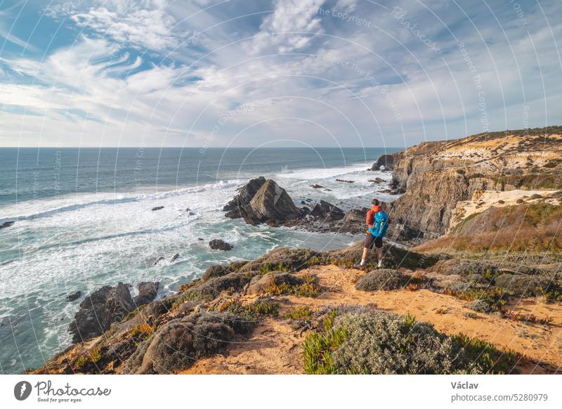 Joyful expression of a young backpacker standing on the edge of a cliff overlooking the Atlantic Ocean in the Odemira region, western Portugal. Wandering along the Fisherman Trail, Rota Vicentina